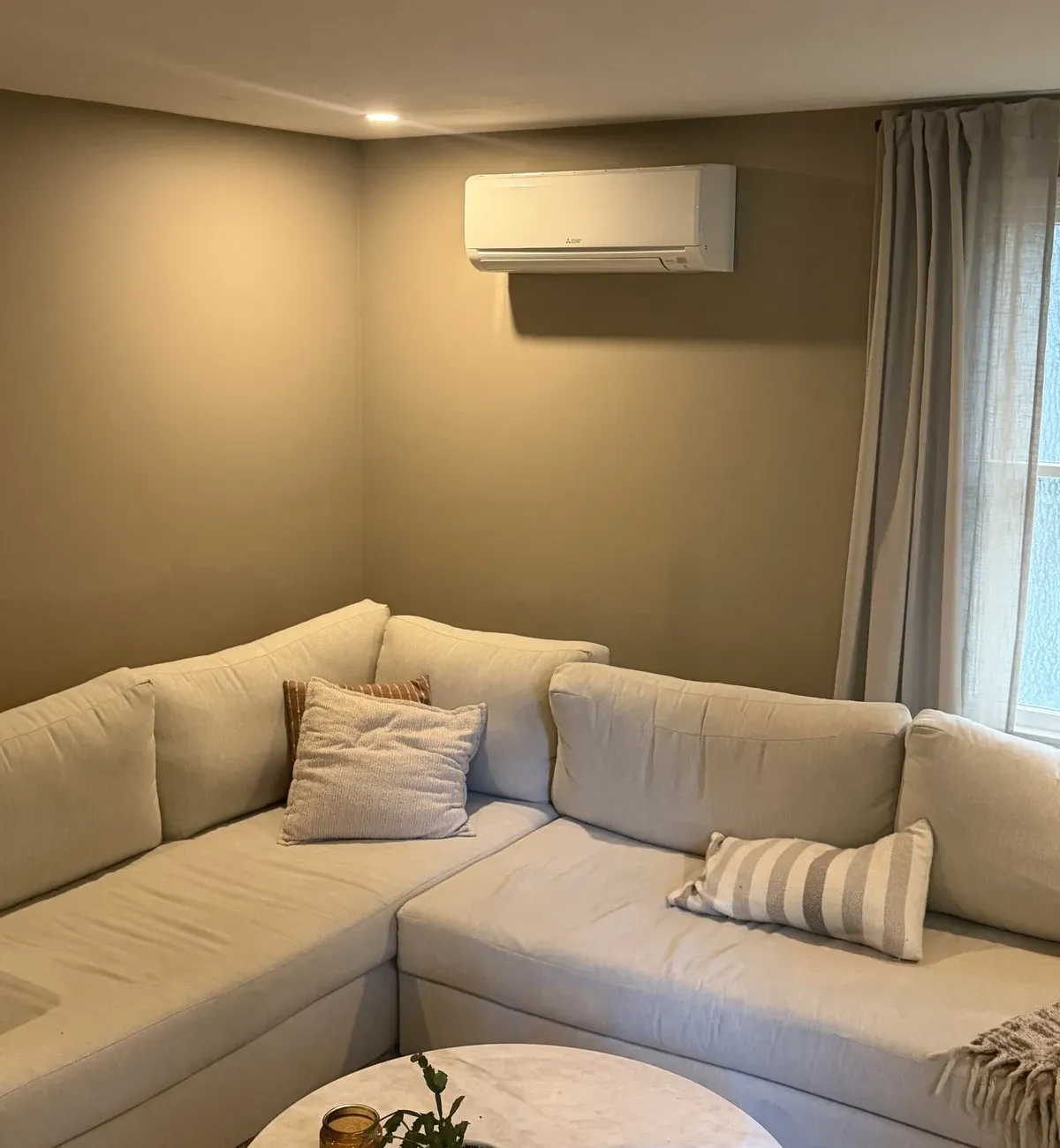 Mitsubishi ductless indoor unit mounted above a neutral sectional sofa beside a curtained window.