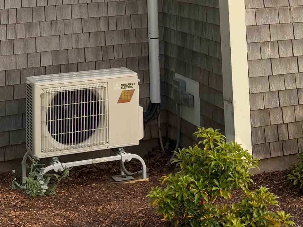 Outdoor Mitsubishi mini-split unit on a metal stand beside cedar shingles, with a covered line set and electrical disconnect.