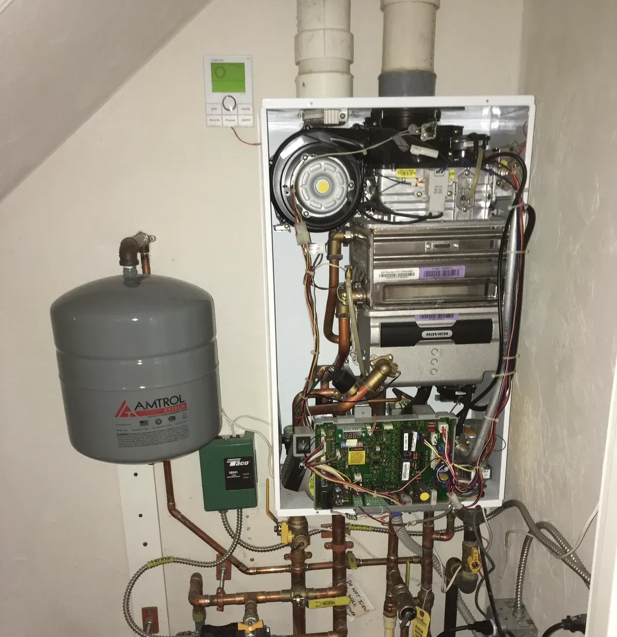 Wall-hung boiler with its access cover open, showing internal components beside an expansion tank and copper piping.